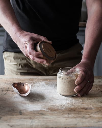 A with a glass jar in his hands with sourdough starter inside, placed on a wooden table.
