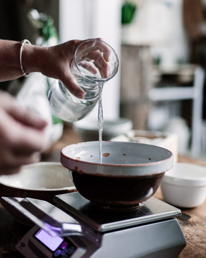 A bowl sits on top of a digital scale as a hand pours water from a jug into the bowl. flour absorption in sourdough