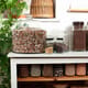 Glass jars of a variety of differnent beans and gains sit on wooden shelves in bright daylight.