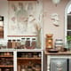A cozy, vintage-inspired kitchen corner featuring a large anatomical illustration of the human digestive system hanging on the wall. Below, a wooden table with jars of various herbs, spices, and seeds. On the right, a ceramic grinder and a decorative white face sculpture. The room features neutral tones with a soft pink wall, dried plants in a glass vase, and antique kitchenware like metal storage containers. A light fixture with a frosted glass globe hangs from the ceiling.