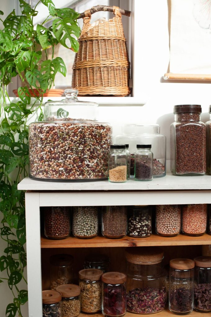 Glass jars of a variety of differnent beans and gains sit on wooden shelves in bright daylight.