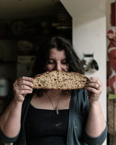A person with dark, curly hair and a nose ring holds up a thick slice of freshly baked bread in front of their face, smiling slightly. They are wearing a black tank top and a cardigan, with flour marks on their skin, suggesting they've been baking. The background shows a cozy kitchen with rustic elements. The bread has a golden-brown crust and visible air pockets in the soft interior.