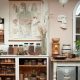 A cozy, vintage-inspired kitchen corner featuring a large anatomical illustration of the human digestive system hanging on the wall. Below, a wooden table with jars of various herbs, spices, and seeds. On the right, a ceramic grinder and a decorative white face sculpture. The room features neutral tones with a soft pink wall, dried plants in a glass vase, and antique kitchenware like metal storage containers. A light fixture with a frosted glass globe hangs from the ceiling.
