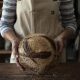A person wearing a beige apron with green straps holds a freshly baked round loaf of bread, displaying a beautiful pattern on the crust. The person is wearing a striped shirt, and the background shows a rustic wooden table, suggesting a warm, home-baked atmosphere. The loaf is dusted with flour, and the hands gently cradle it as the bread's textured surface is prominently featured.
