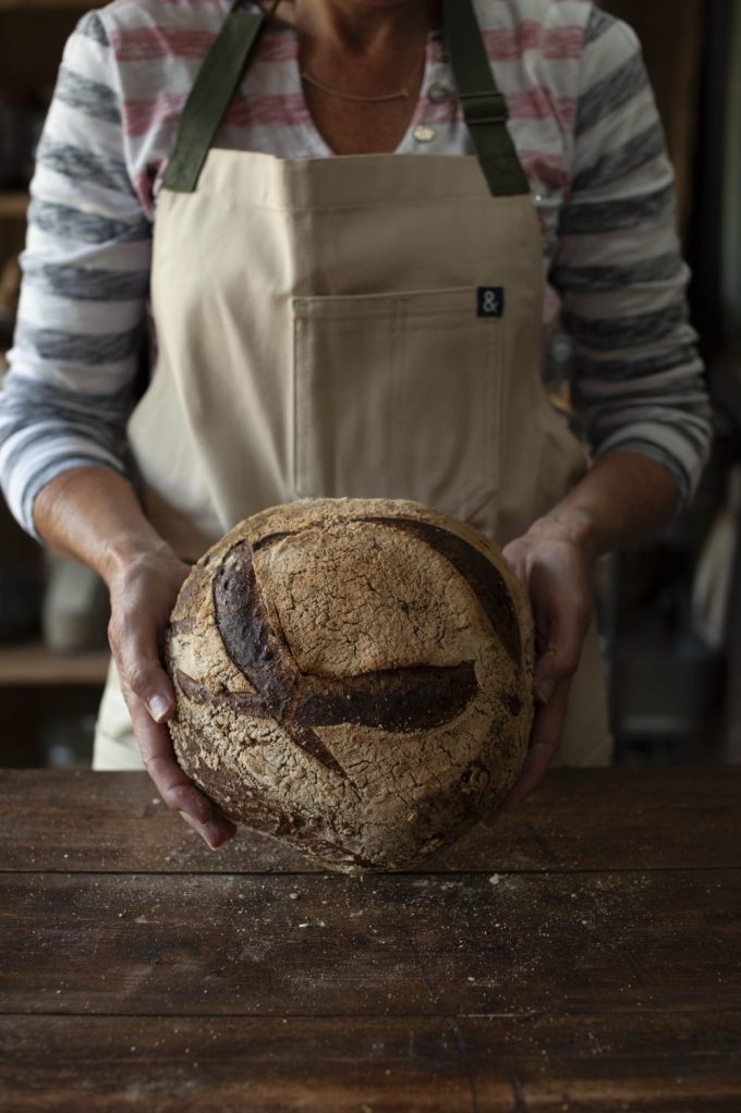 A person wearing a beige apron with green straps holds a freshly baked round loaf of bread, displaying a beautiful pattern on the crust. The person is wearing a striped shirt, and the background shows a rustic wooden table, suggesting a warm, home-baked atmosphere. The loaf is dusted with flour, and the hands gently cradle it as the bread's textured surface is prominently featured.